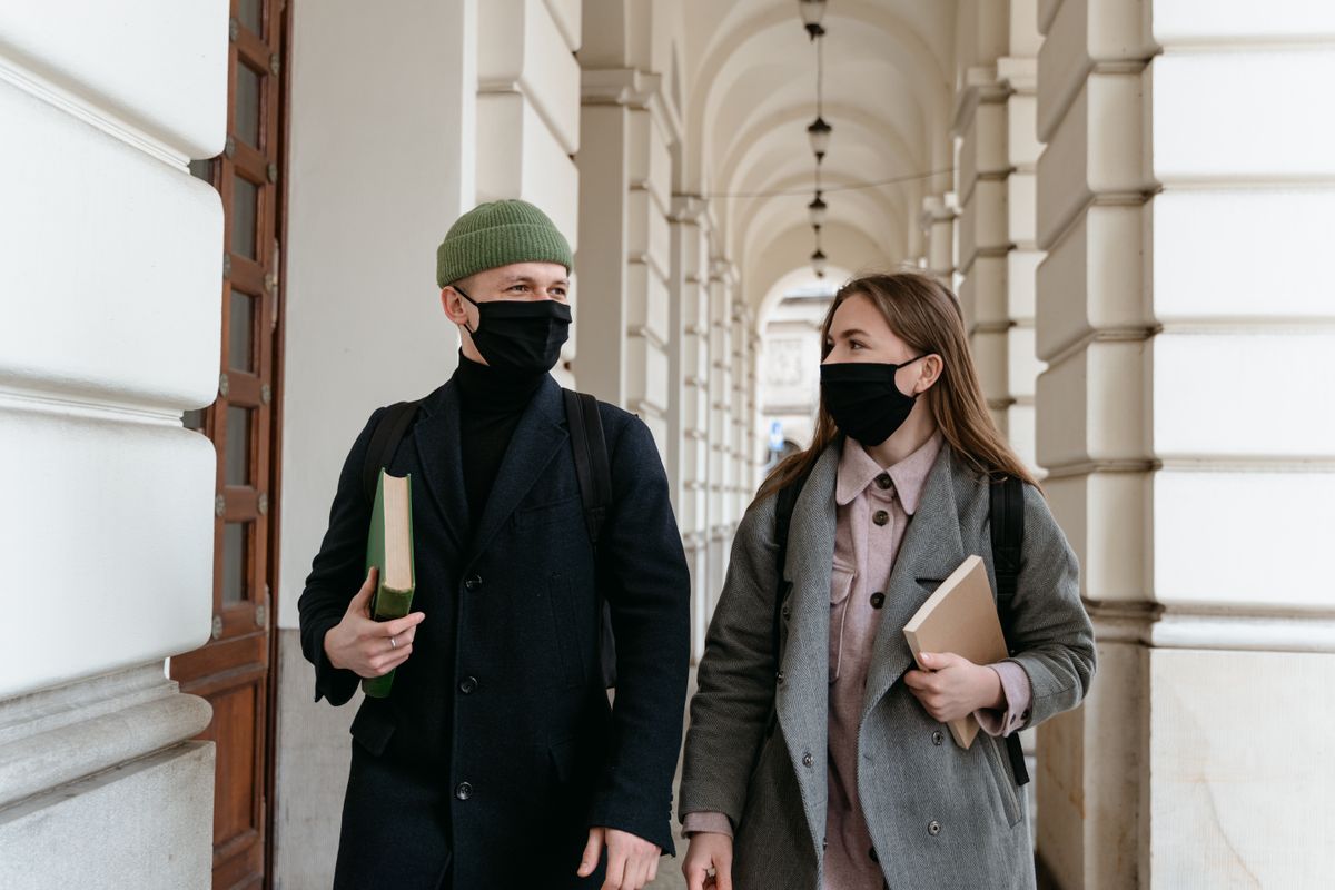 Two students in face masks holding books walking through a historic hallway, showcasing new normal lifestyle.