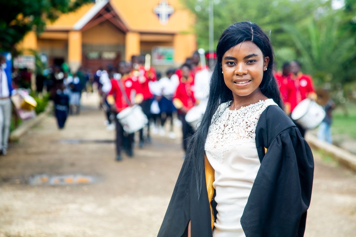 A young woman in a graduation gown smiles outdoors with a marching band in Lusaka, Zambia.