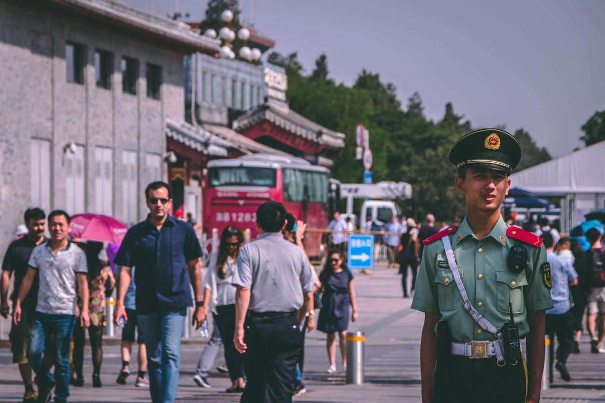 A Chinese police officer stands on a bustling street with people walking by on a sunny day.