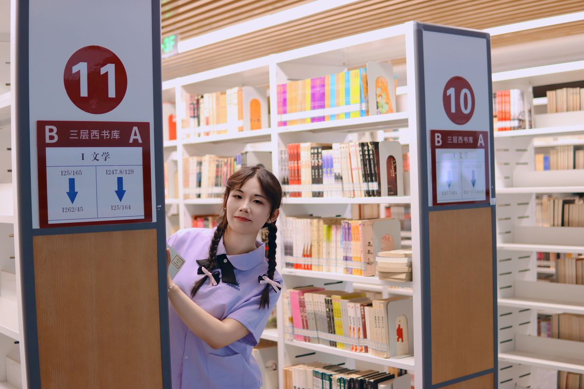 A woman in a library aisle, surrounded by colorful books and signs, looking cheerful.
