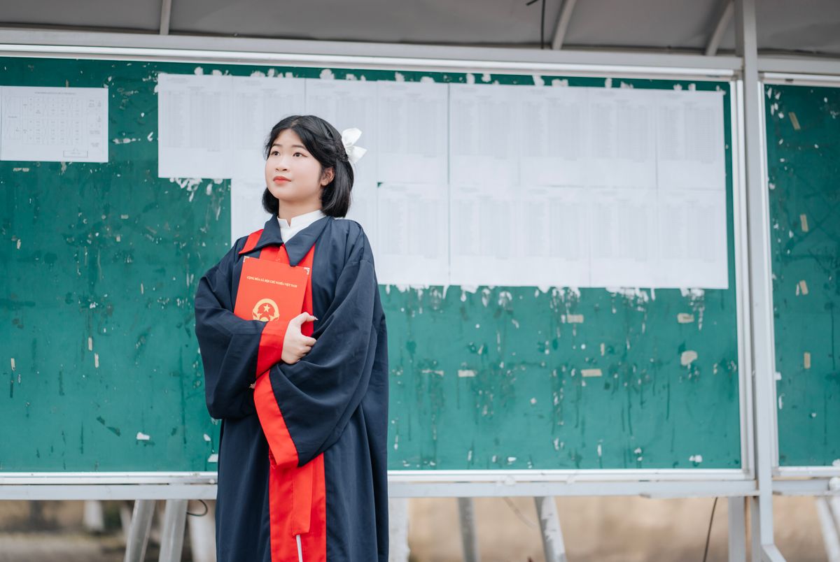 Young graduate in academic gown holding diploma, standing in front of outdoor bulletin board.