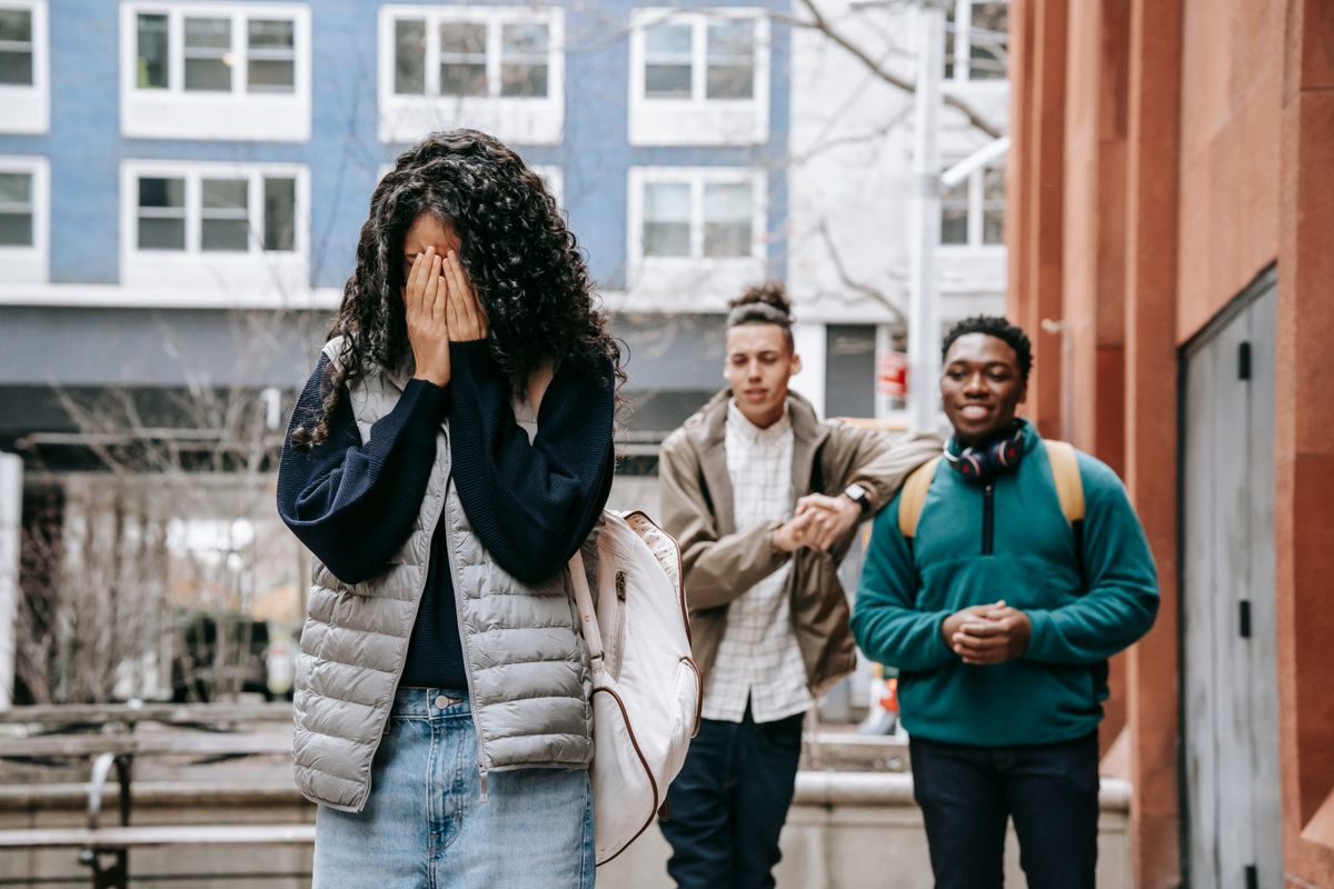 Unrecognizable young female student covering face with hands while being bullied by multiethnic male classmates on street